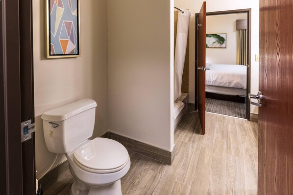 Un baño de hotel con inodoro, ducha y una cama visible a través de una puerta abierta.