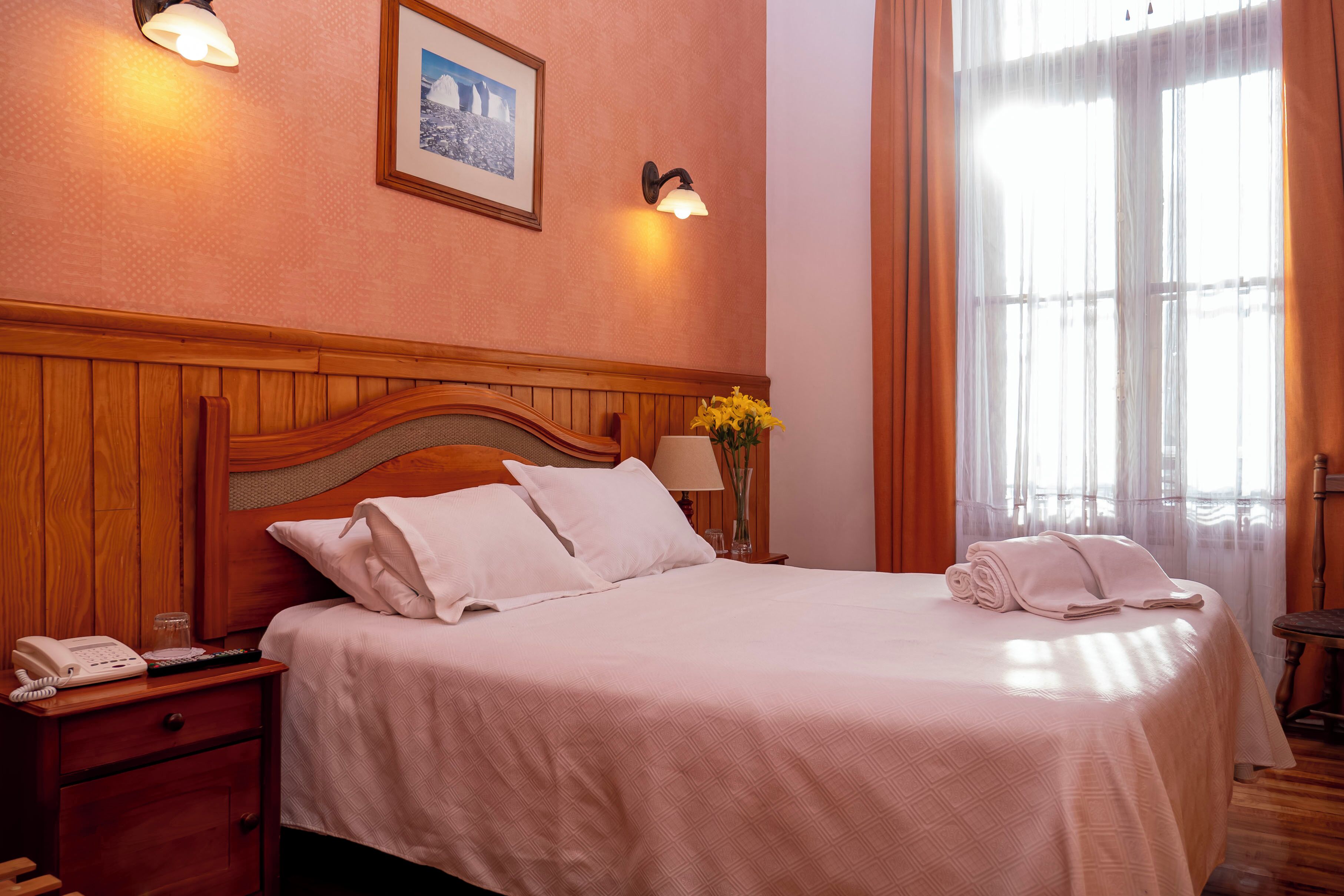 A neatly made bed with white linens, a bedside lamp, a framed picture on the wall, and a window with curtains.