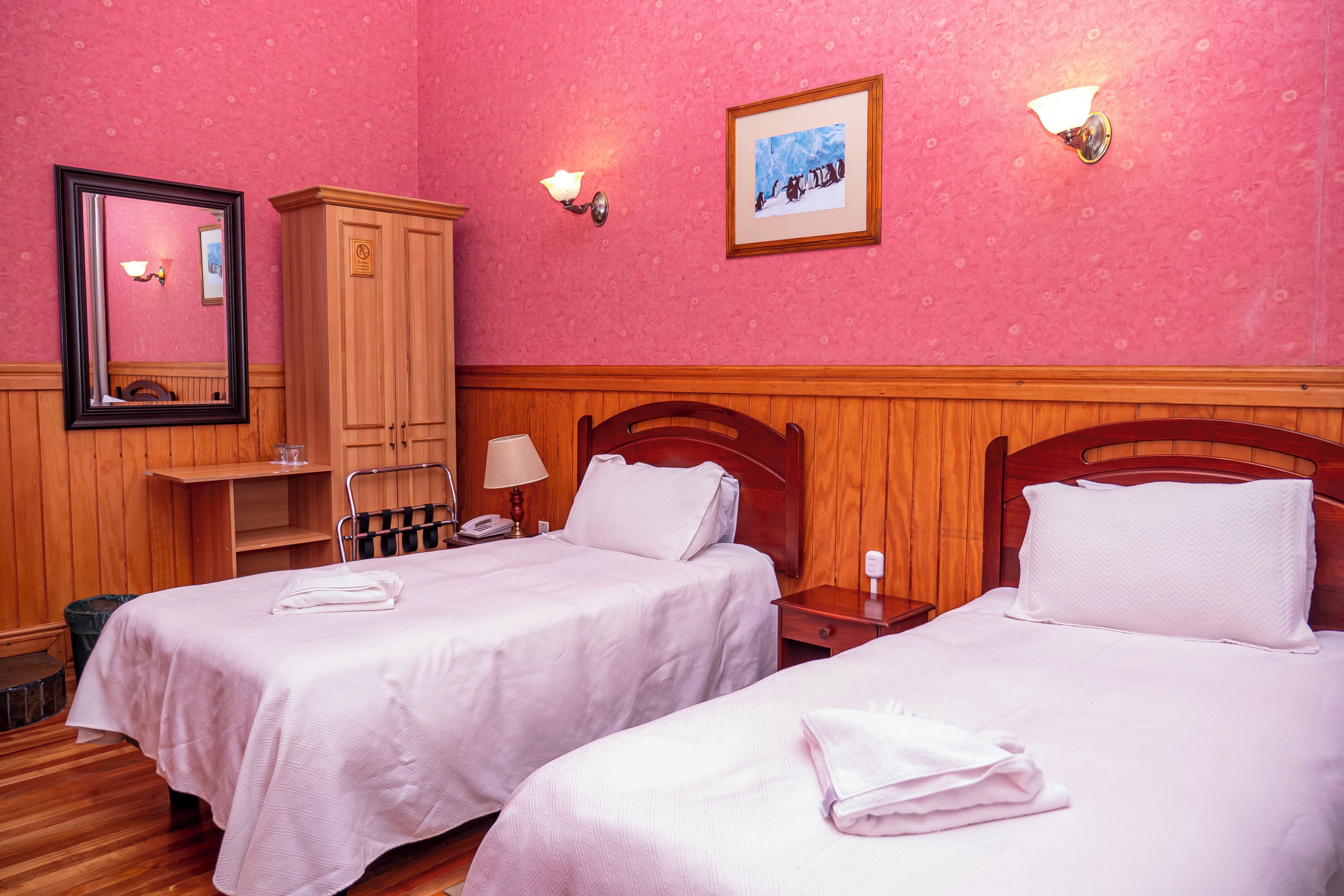 A hotel room with two beds, wooden headboards, a wooden wardrobe, a mirror, and a framed picture on the wall.
