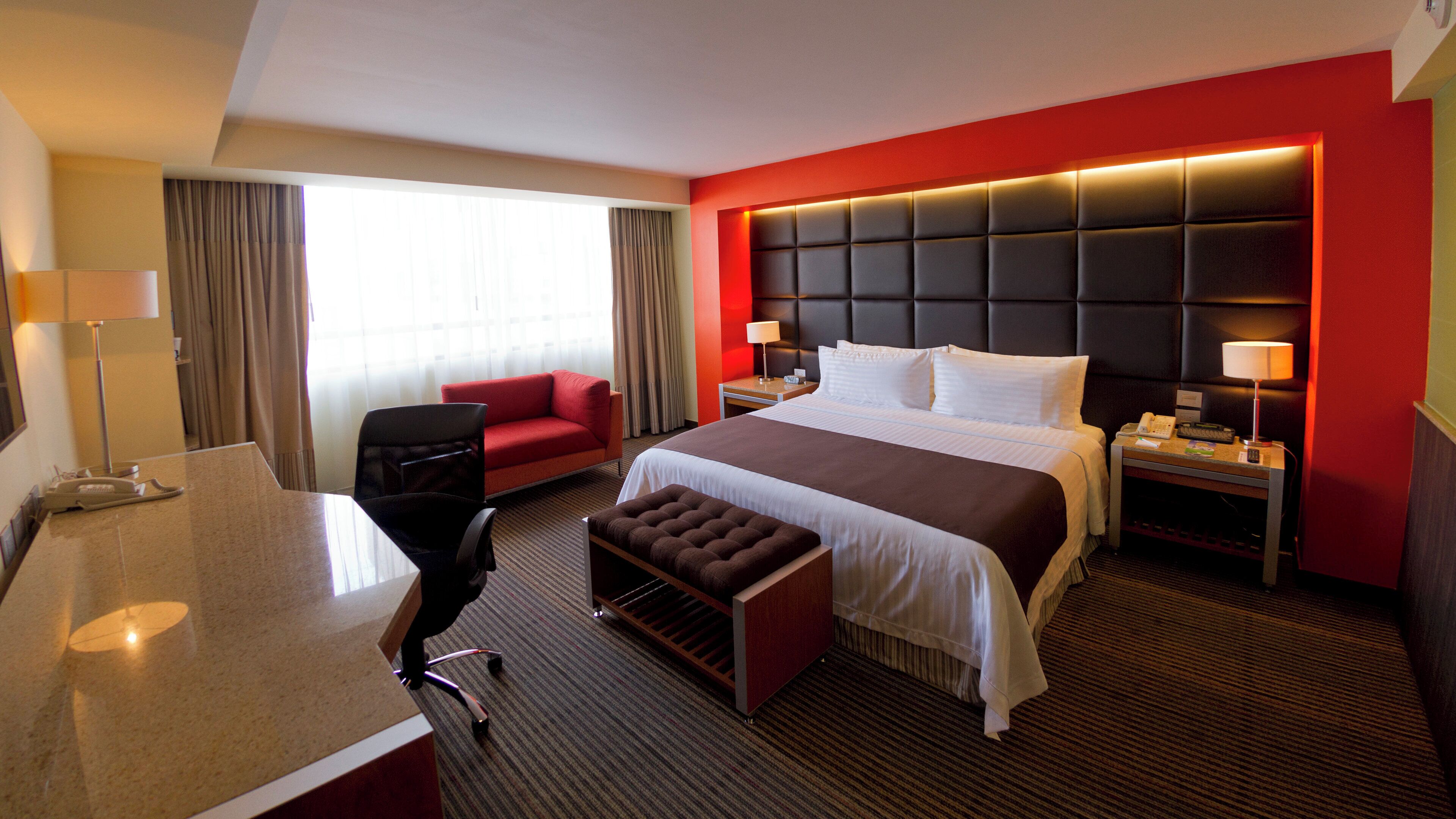 Habitación de hotel con cama, escritorio, silla, mesita y mesita de noche con lámpara.