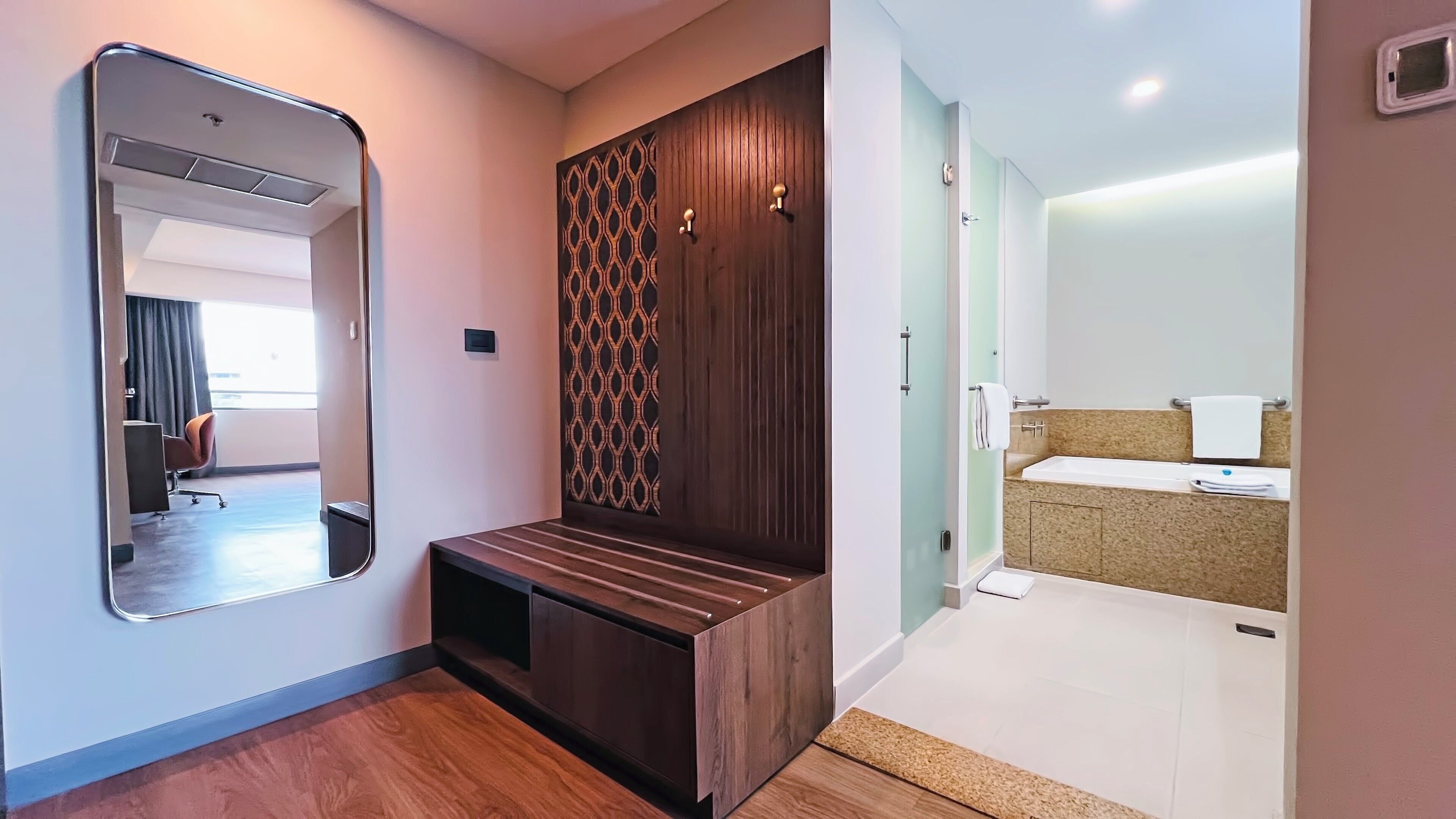 Une chambre d’hôtel avec un banc en bois, un grand miroir, un mur à motifs et une salle de bain équipée d’un lavabo et d’un porte-serviettes.