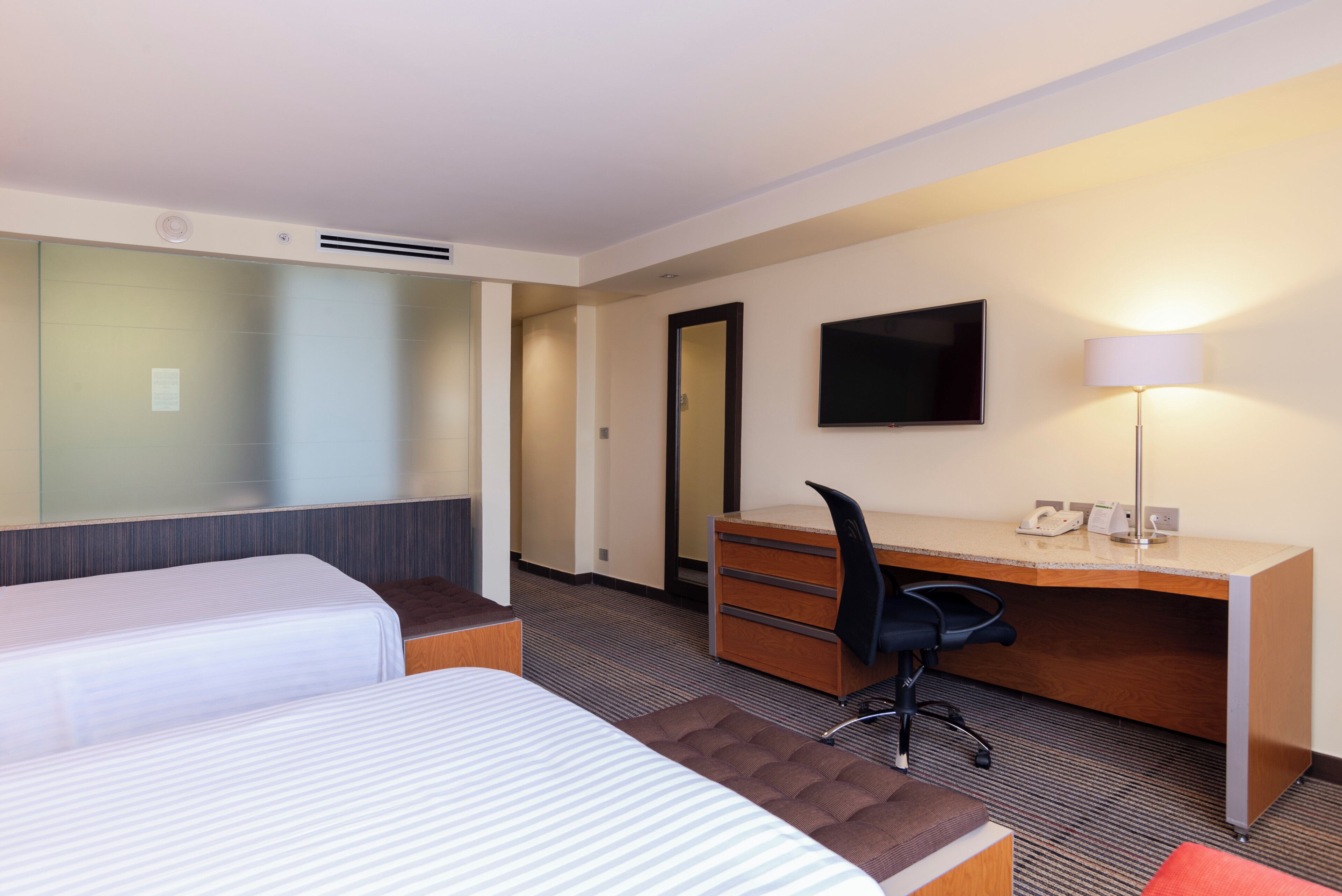 A hotel room with two beds, a desk, a chair, a flat-screen TV, and a lamp.