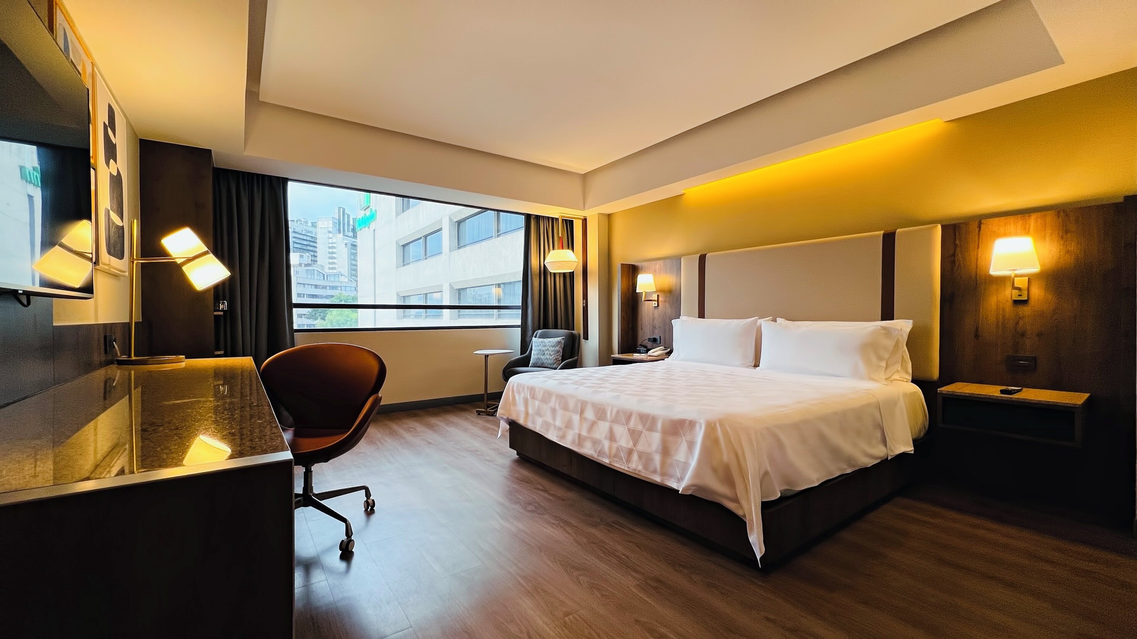 A hotel room with a large bed, a desk with a chair, a small table, and a window with a view of buildings.