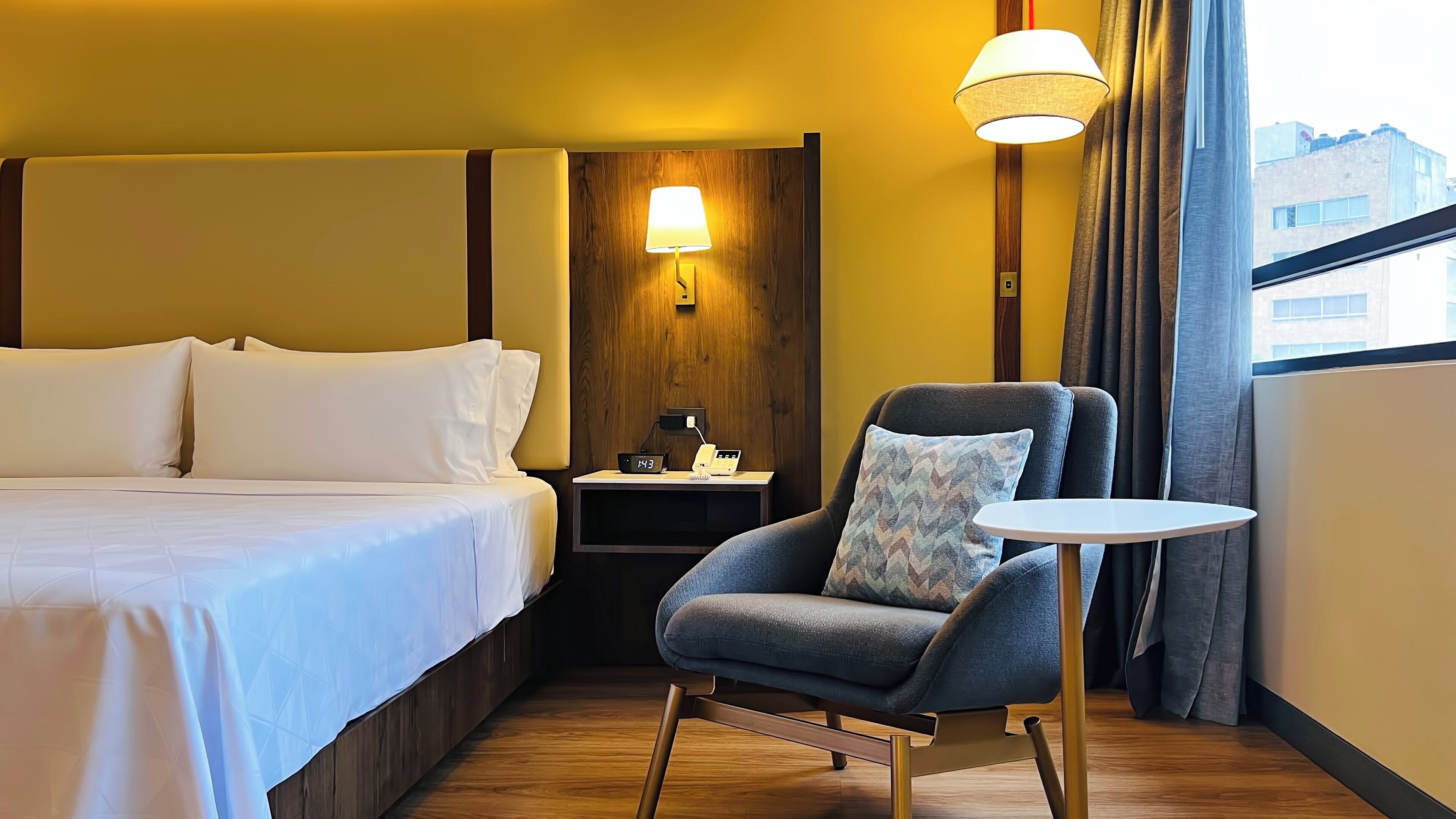 Une chambre d’hôtel avec un lit, une chaise, une petite table, une table de chevet, une lampe et une fenêtre avec des rideaux.