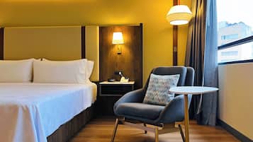 Habitación de hotel con cama, silla, mesita, mesita de noche, lámpara y ventana con cortinas.