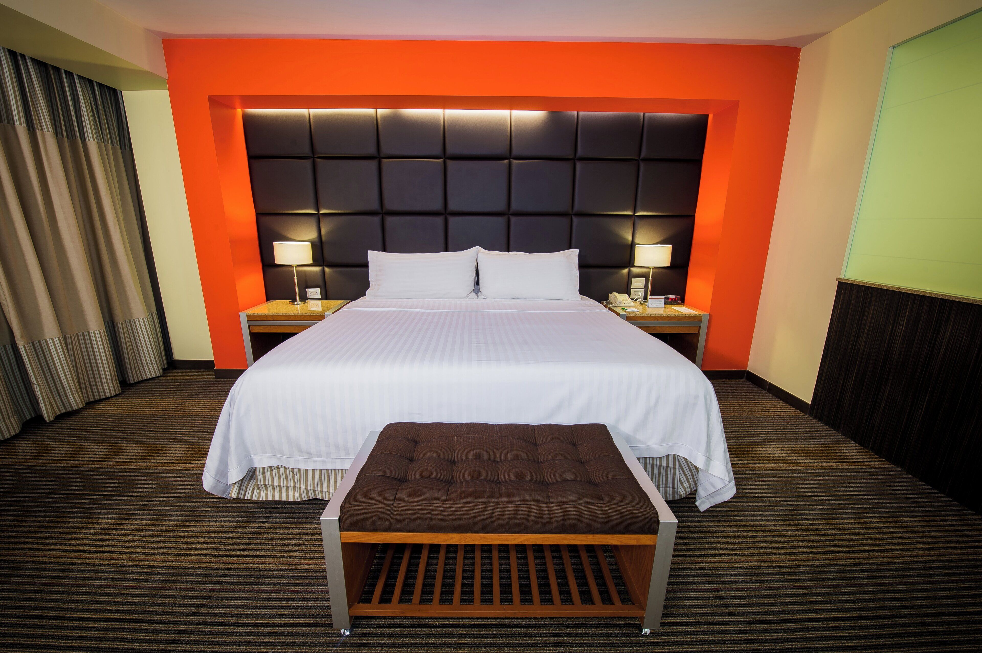A hotel room with a large bed, two bedside tables, a nightstand, a bench, and a headboard with built-in lighting.