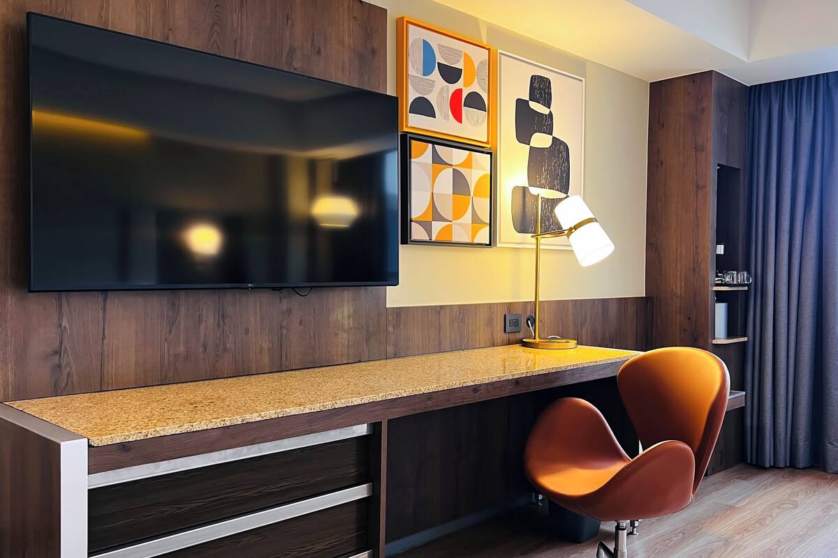 A modern hotel room with a flat-screen TV mounted on the wall, a desk with a lamp, and a comfortable orange chair.