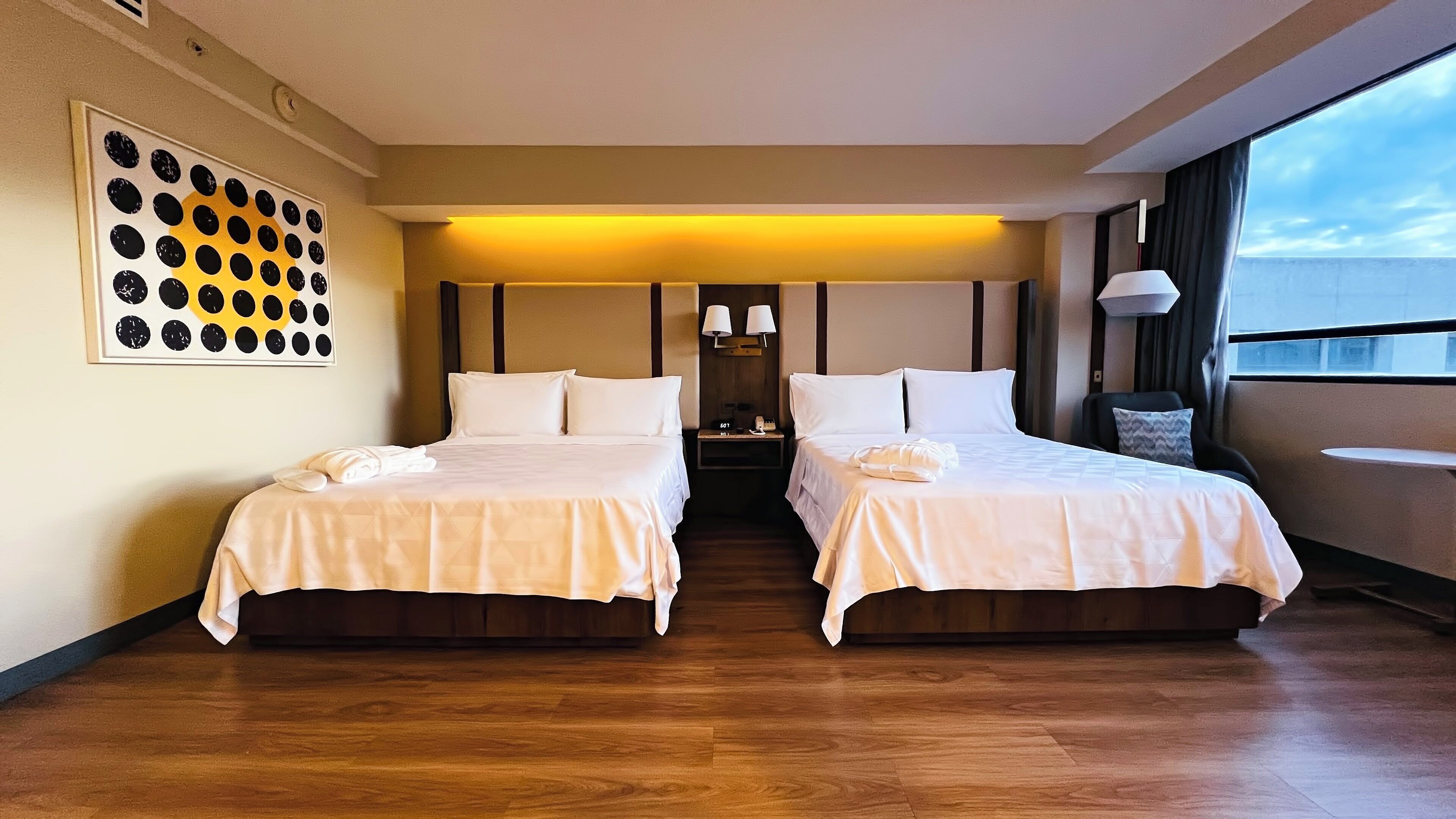 Habitación de hotel con dos camas, una mesita de noche, un reloj de pared y una ventana con vistas.