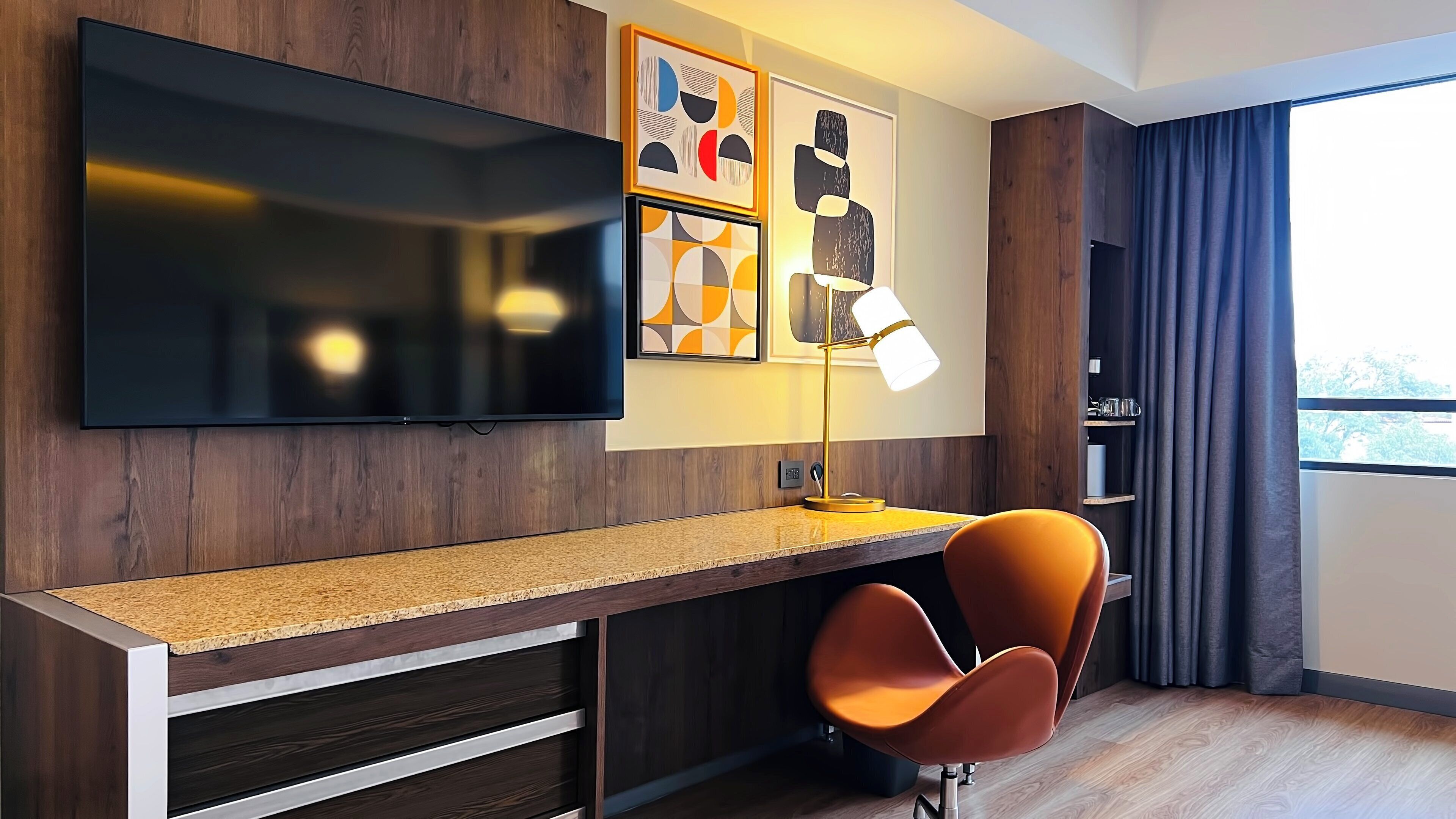 Une chambre d’hôtel moderne dotée d’un téléviseur à écran plat fixé au mur, d’un bureau avec une lampe et d’un fauteuil confortable de couleur orange.