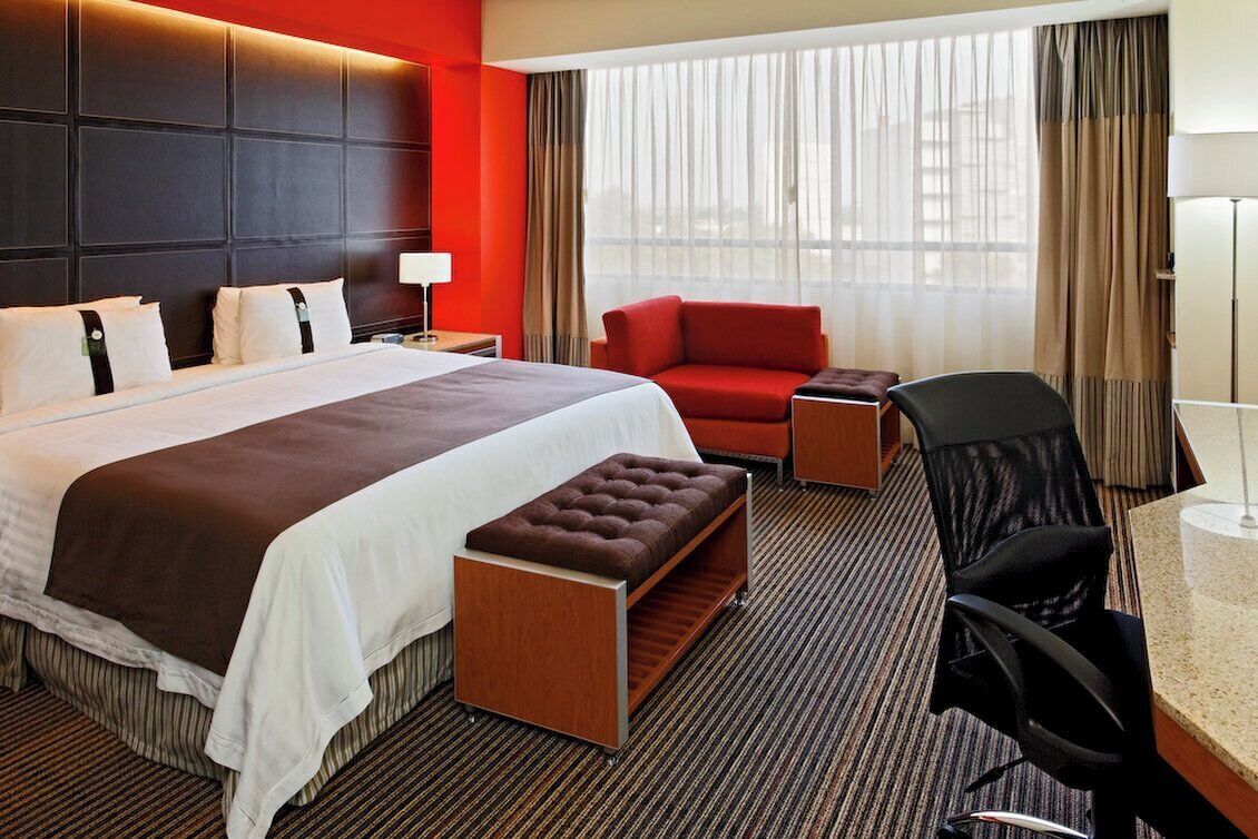 A hotel room with a large bed, a red armchair, a desk with a chair, and a window with curtains.