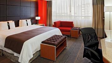 A hotel room with a large bed, a red armchair, a desk with a chair, and a window with curtains.
