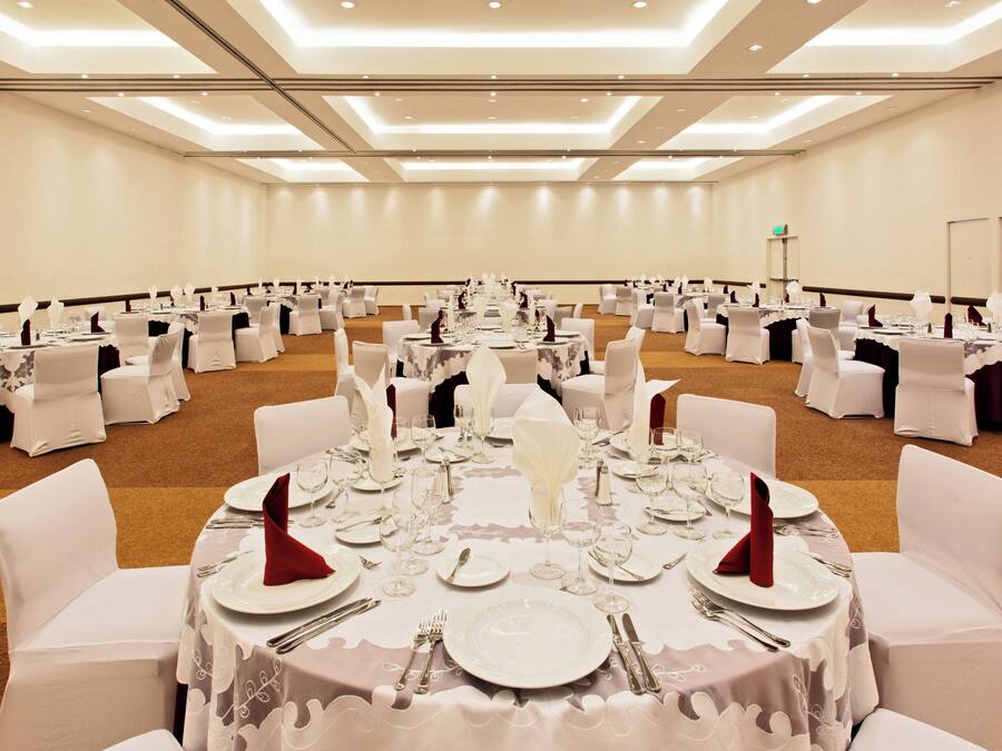 Un salón de banquetes con mesas redondas dispuestas para un evento formal, con manteles blancos, cubiertos y cristalería.