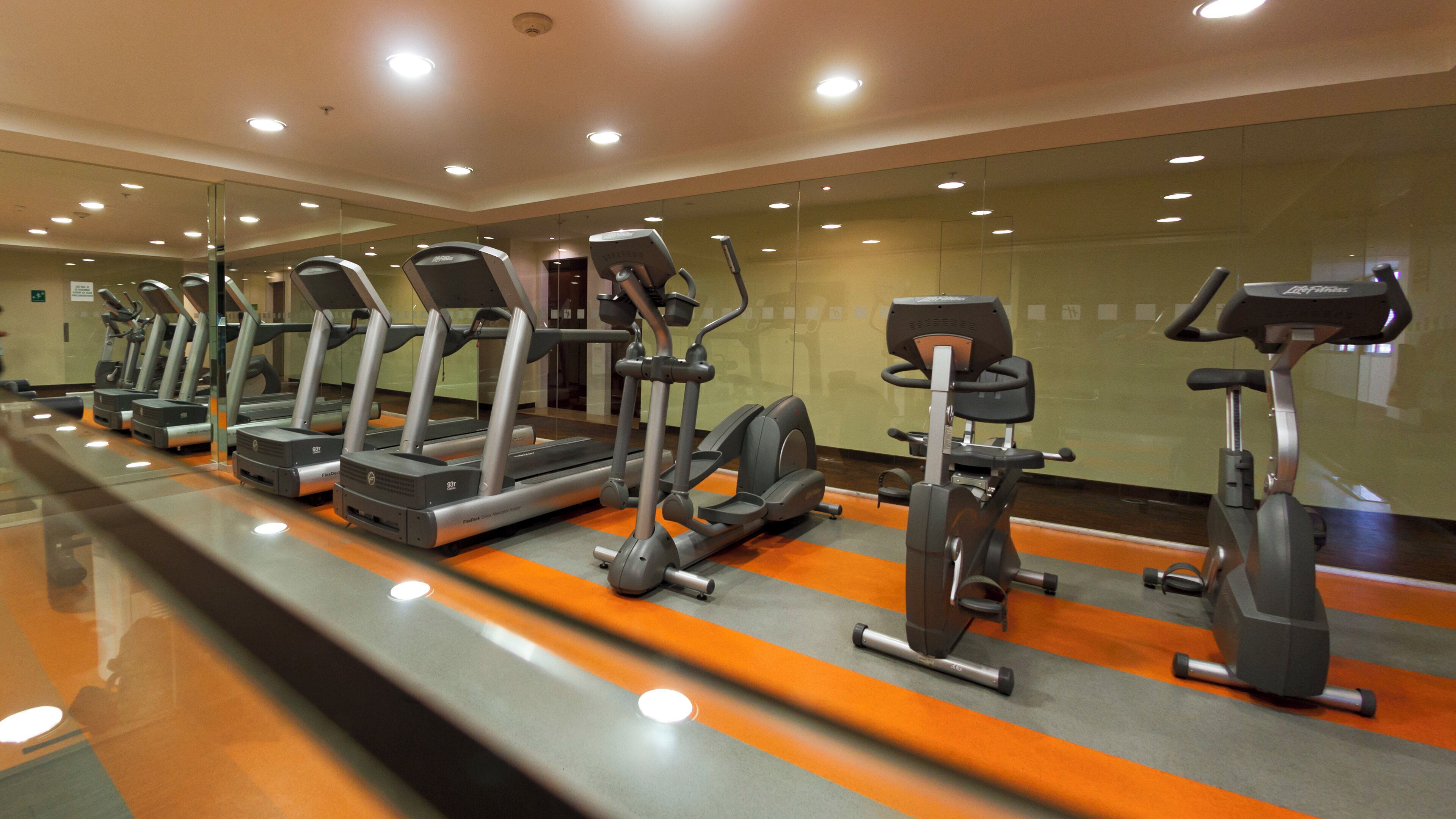 A row of stationary bikes and treadmills in a gym with mirrored walls and a polished floor.