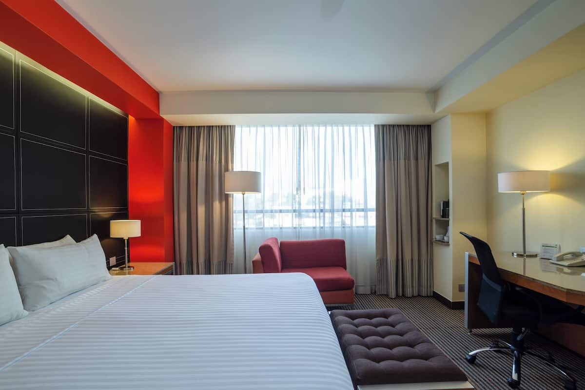 A hotel room with a large bed, a red armchair, a desk with a chair, and a window with curtains.
