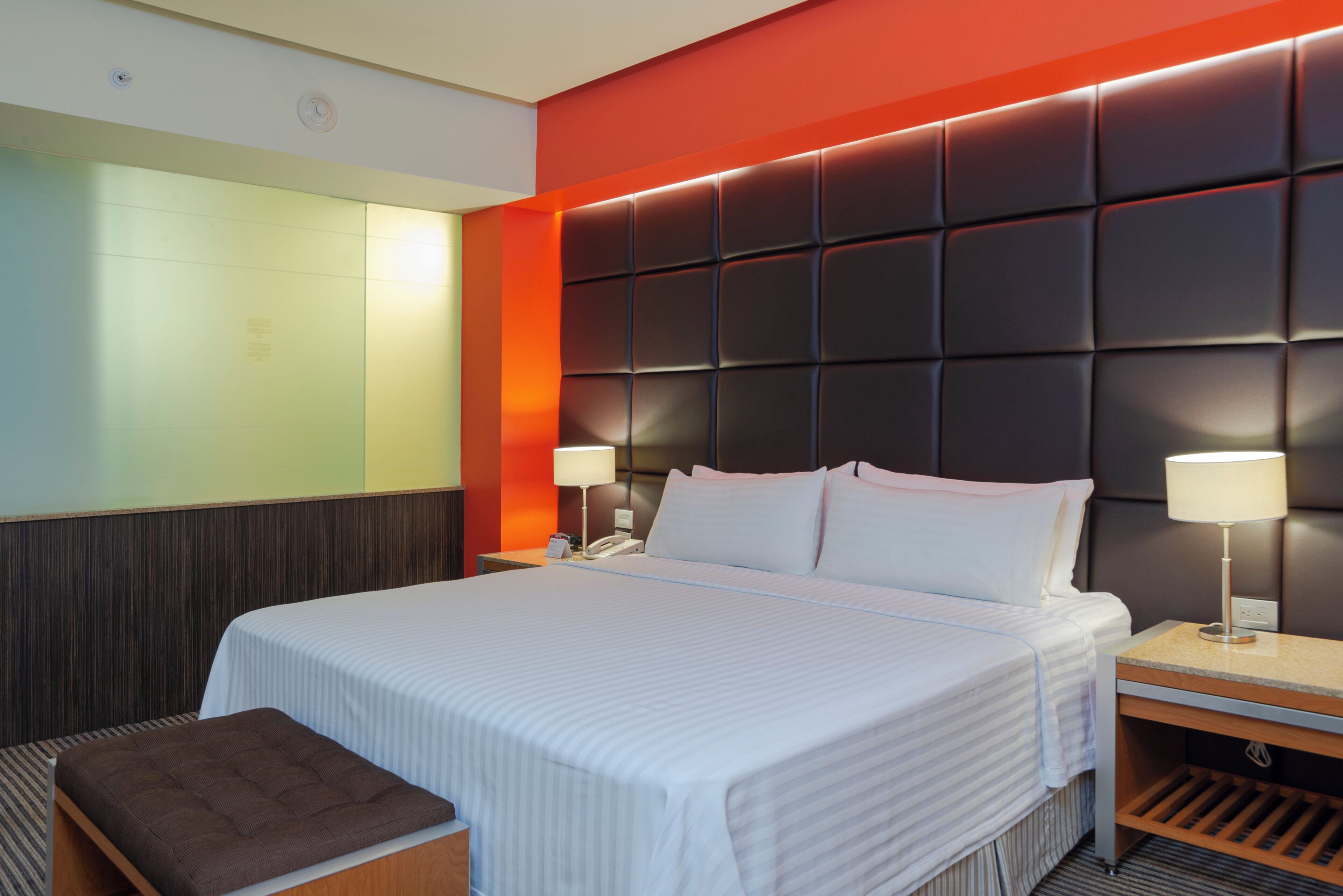 A hotel room with a large bed, bedside lamps, a wooden bench, and a modern headboard.
