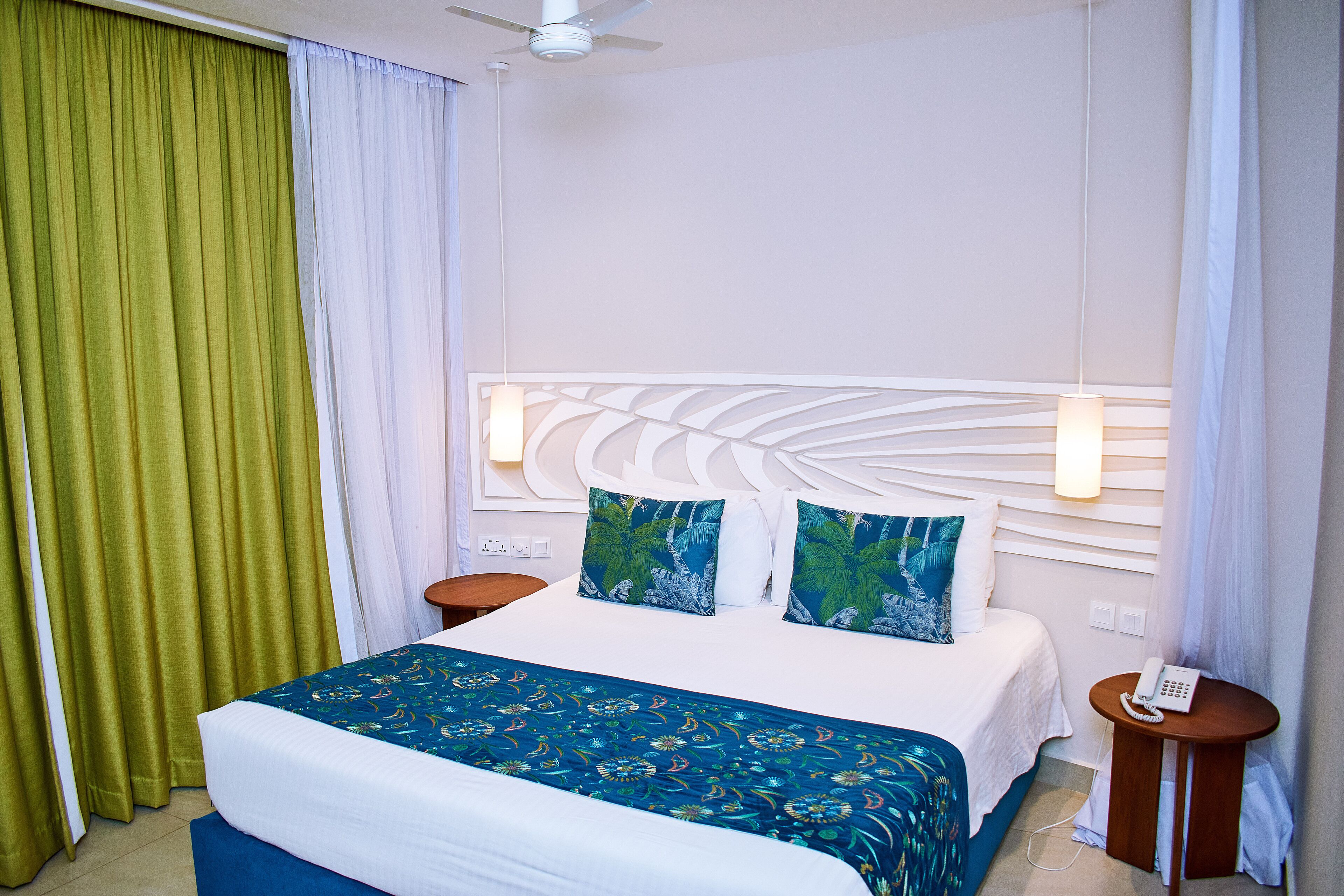 Une chambre d’hôtel avec un lit, deux tables de chevet, un ventilateur de plafond et une petite table de nuit avec un téléphone.