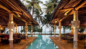 A poolside area with a clear view of the ocean, palm trees, and a thatched roof structure. - Neptune Village Beach Resort & Spa All Inclusive (Diani Beach)