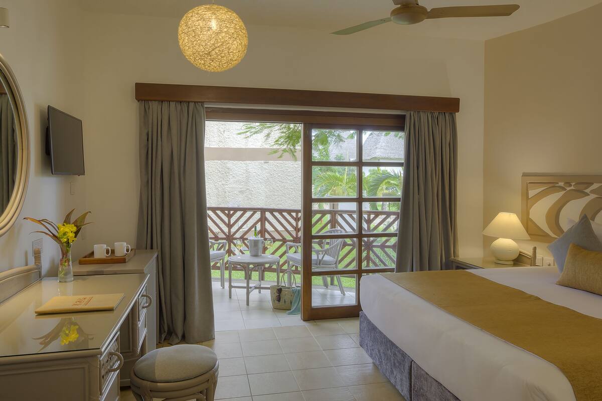A hotel room with a bed, a desk with a computer, a chair, a table with a chair, a balcony with a table and chairs, and a view of greenery outside.
