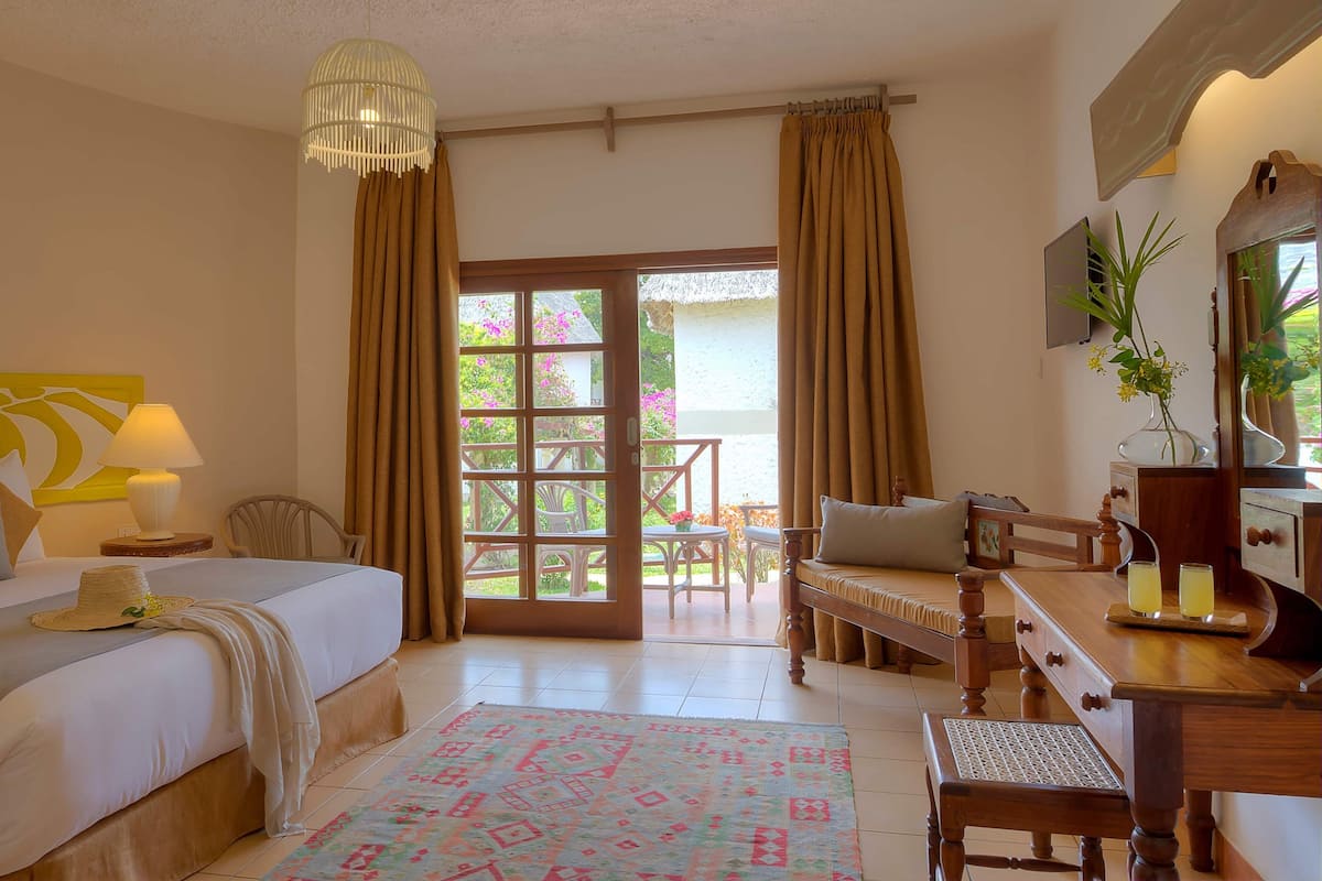A hotel room with a bed, a desk, a chair, a lamp, a rug, and a view of the outside.