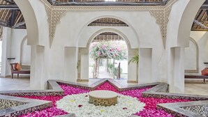 Un vestíbulo de hotel con una zona de estar circular central rodeada por un suelo con mosaicos, pétalos de flores blancos y rosa, y vistas a un jardín a través de un arco.