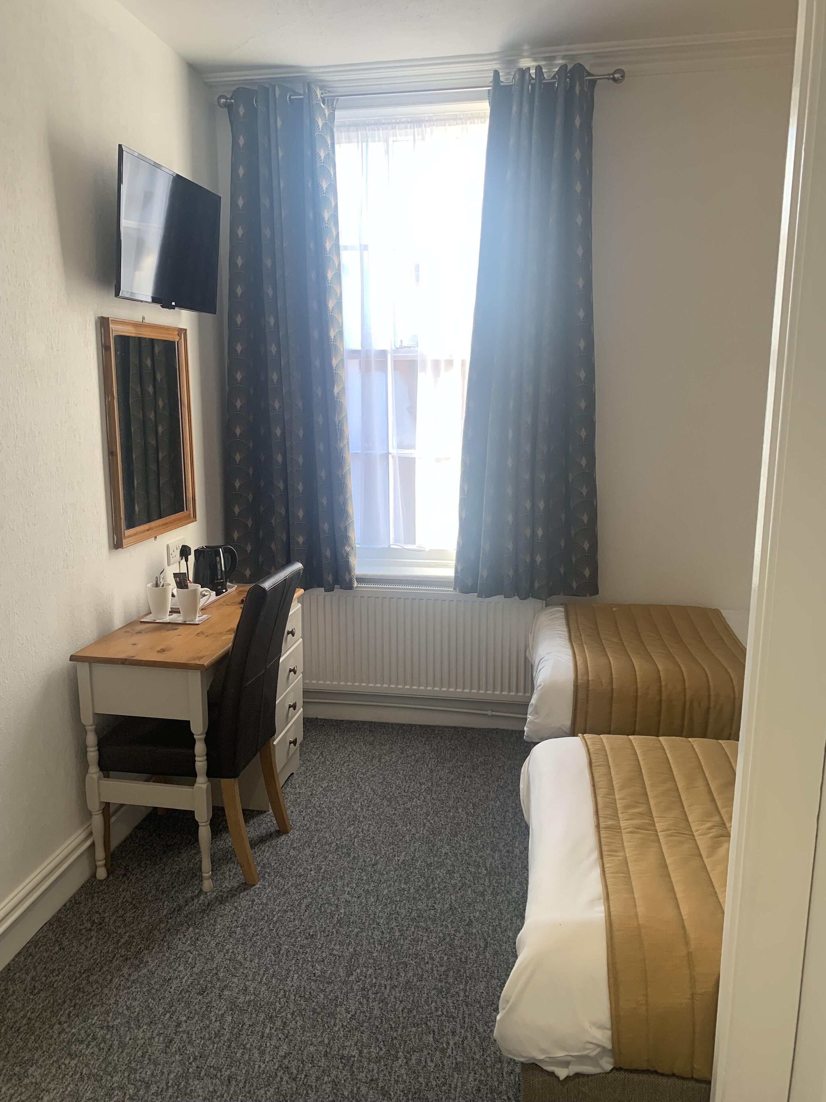A bedroom with a bed, a desk with a chair, a window with curtains, a radiator, and a television mounted on the wall.