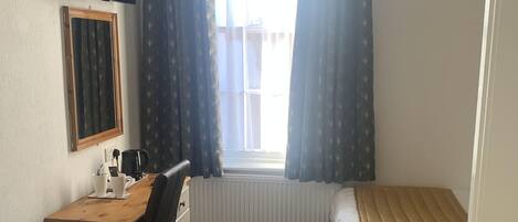 Ein Schlafzimmer mit einem Bett, einem Schreibtisch mit Stuhl, einem Fenster mit Vorhängen, einem Heizkörper und einem an der Wand befestigten Fernseher.