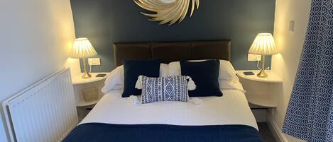 Ein ordentlich bezogenes Bett mit blauer Decke und Kissen, zwei Nachttischlampen, einem dekorativen Wandspiegel und einem gemusterten Vorhang.