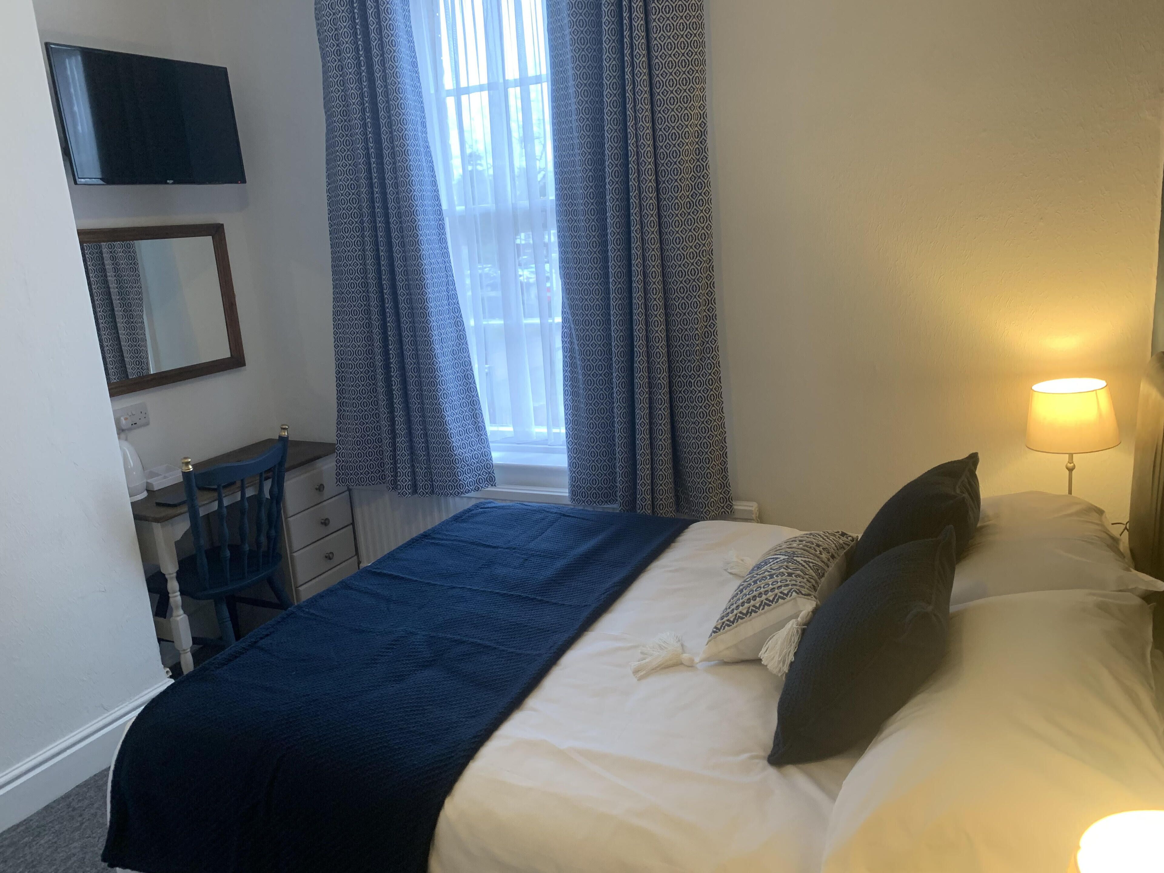 Een slaapkamer met een bed, een bureau, een stoel, een lamp en een raam met gordijnen met een patroon.