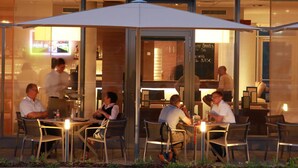 Un espacio de comedor al aire libre con mesas y sillas, un cerramiento de vidrio y vistas al restaurante interior.