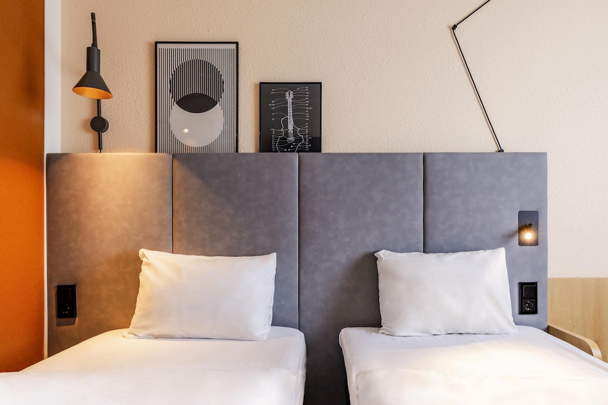 A hotel room with two beds, a modern headboard, a wall-mounted lamp, and a framed anatomical diagram.
