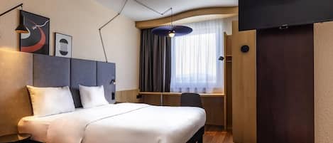 A hotel room with a bed, a desk, a chair, a TV, and a window with curtains.
