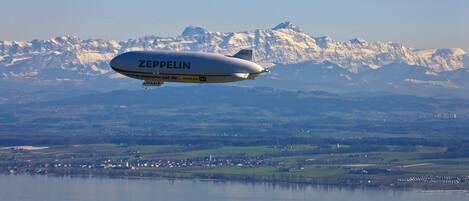 Un dirigible Zeppelin volando sobre un lago, con un paisaje montañoso al fondo.