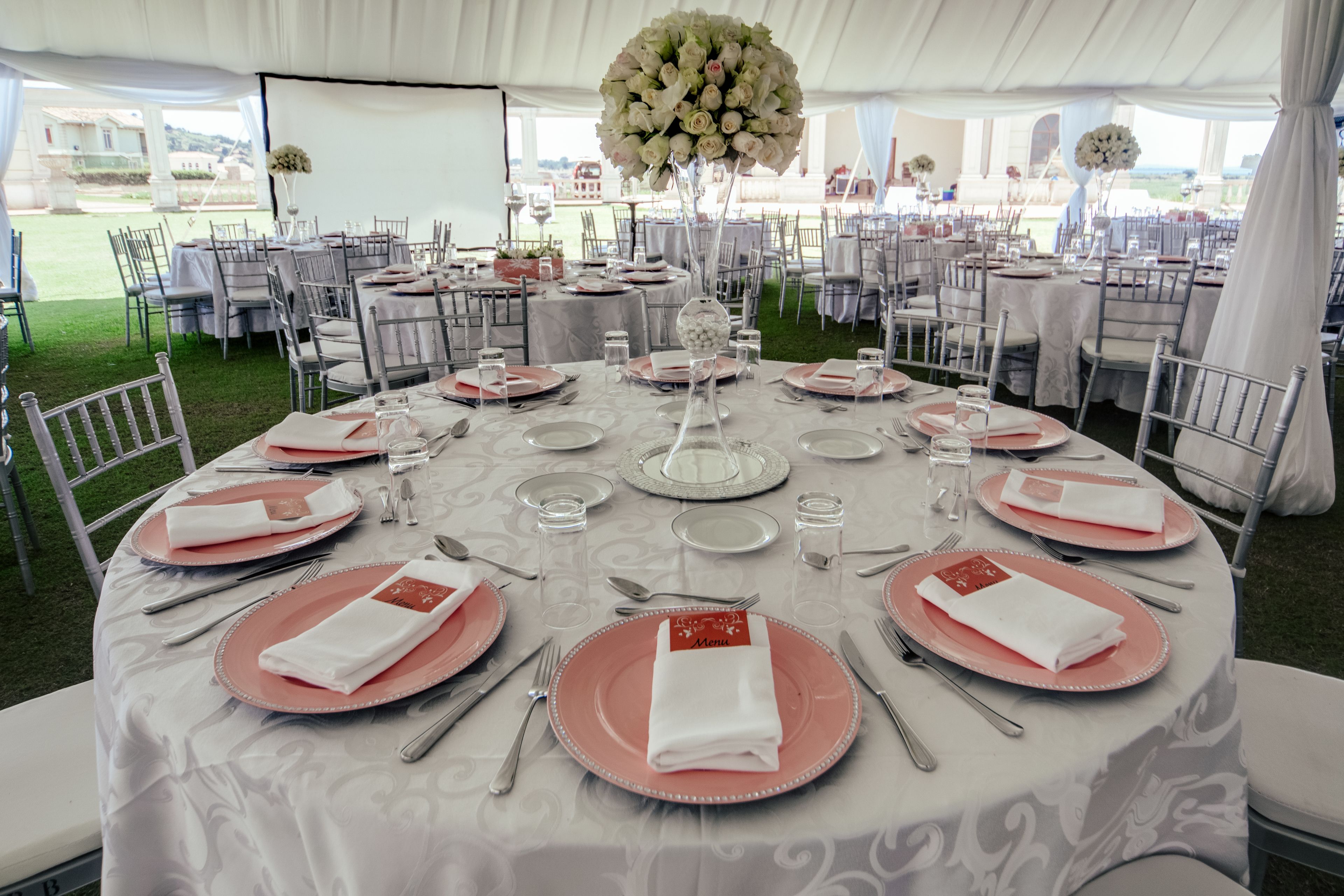 A tented outdoor event space with round tables set for dining, featuring white tablecloths and pink plates.