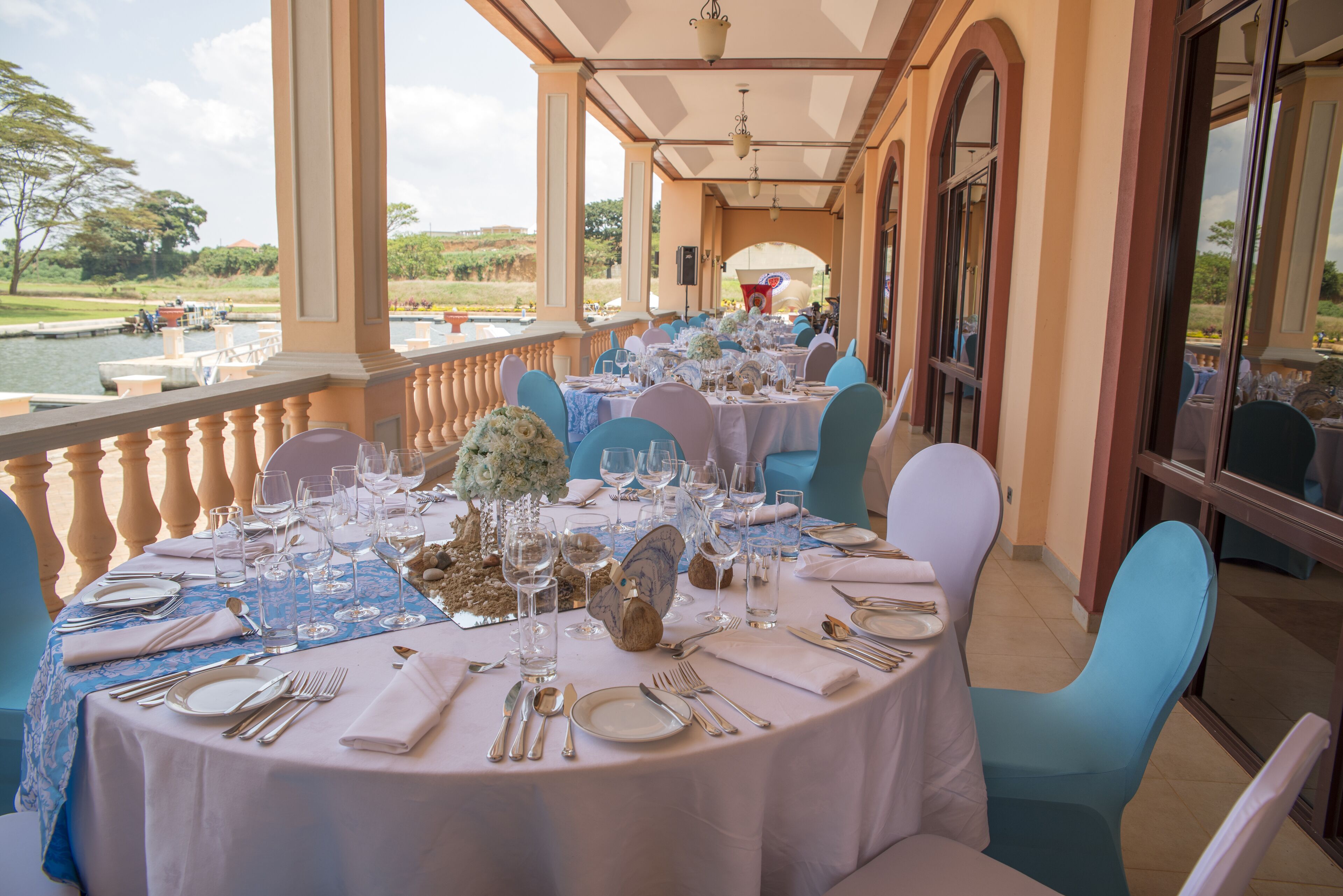 A dining area with round tables set for a formal meal, overlooking a body of water and greenery.
