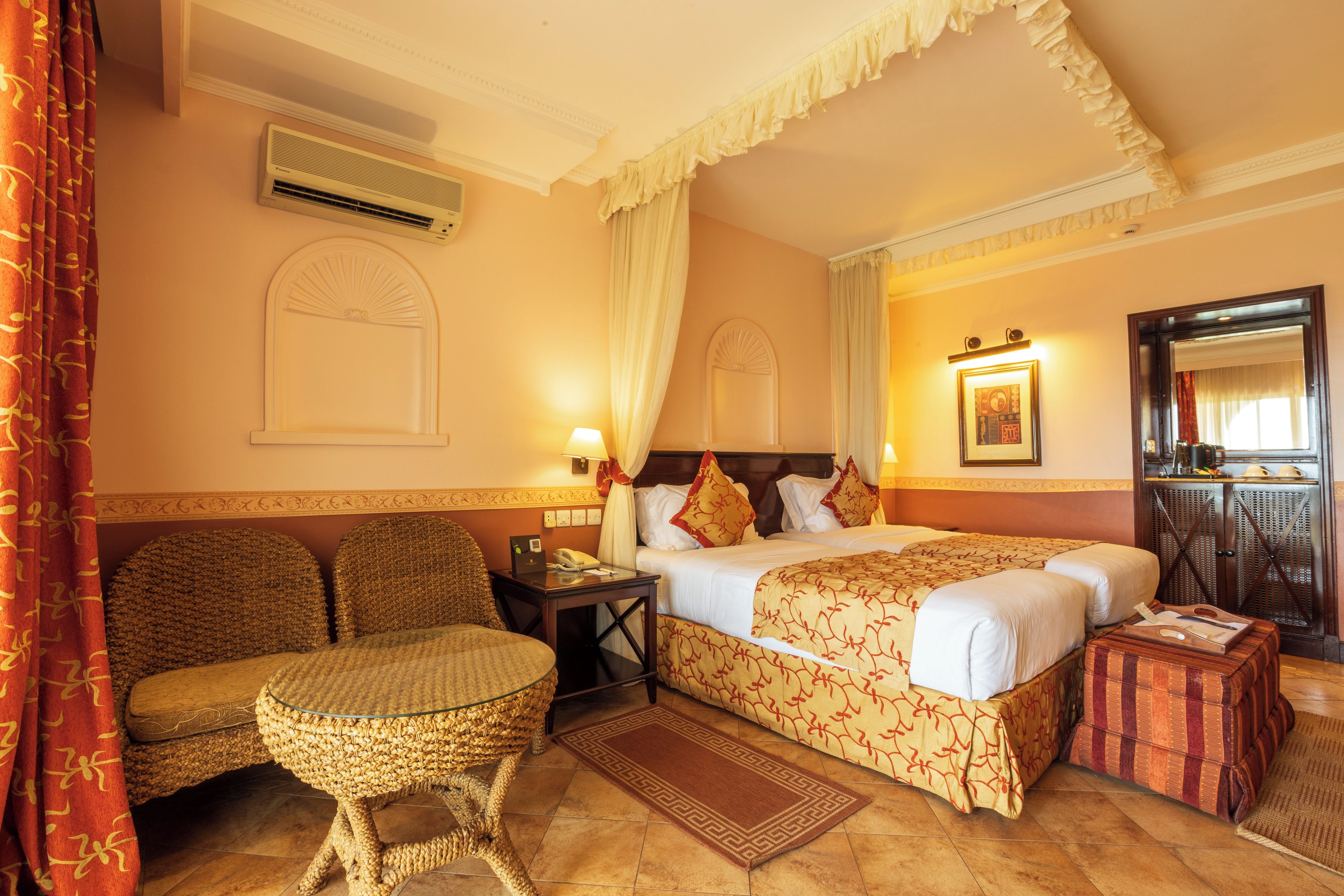 A hotel room with a bed, a sitting area with a chair, a small table, a bedside table with a lamp, and a mirror on the wall.