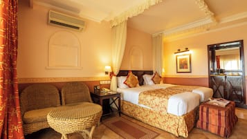 A hotel room with a bed, a sitting area with a chair, a small table, a bedside table with a lamp, and a mirror on the wall.