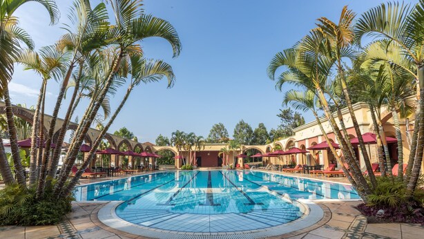 A large outdoor swimming pool surrounded by palm trees and red umbrellas, with a tiled walkway and a building in the background. - Lake Victoria Serena Golf Resort & Spa (Kampala)