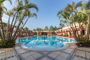 A large outdoor swimming pool surrounded by palm trees and red umbrellas, with a tiled walkway and a building in the background. - Lake Victoria Serena Golf Resort & Spa (Kampala)