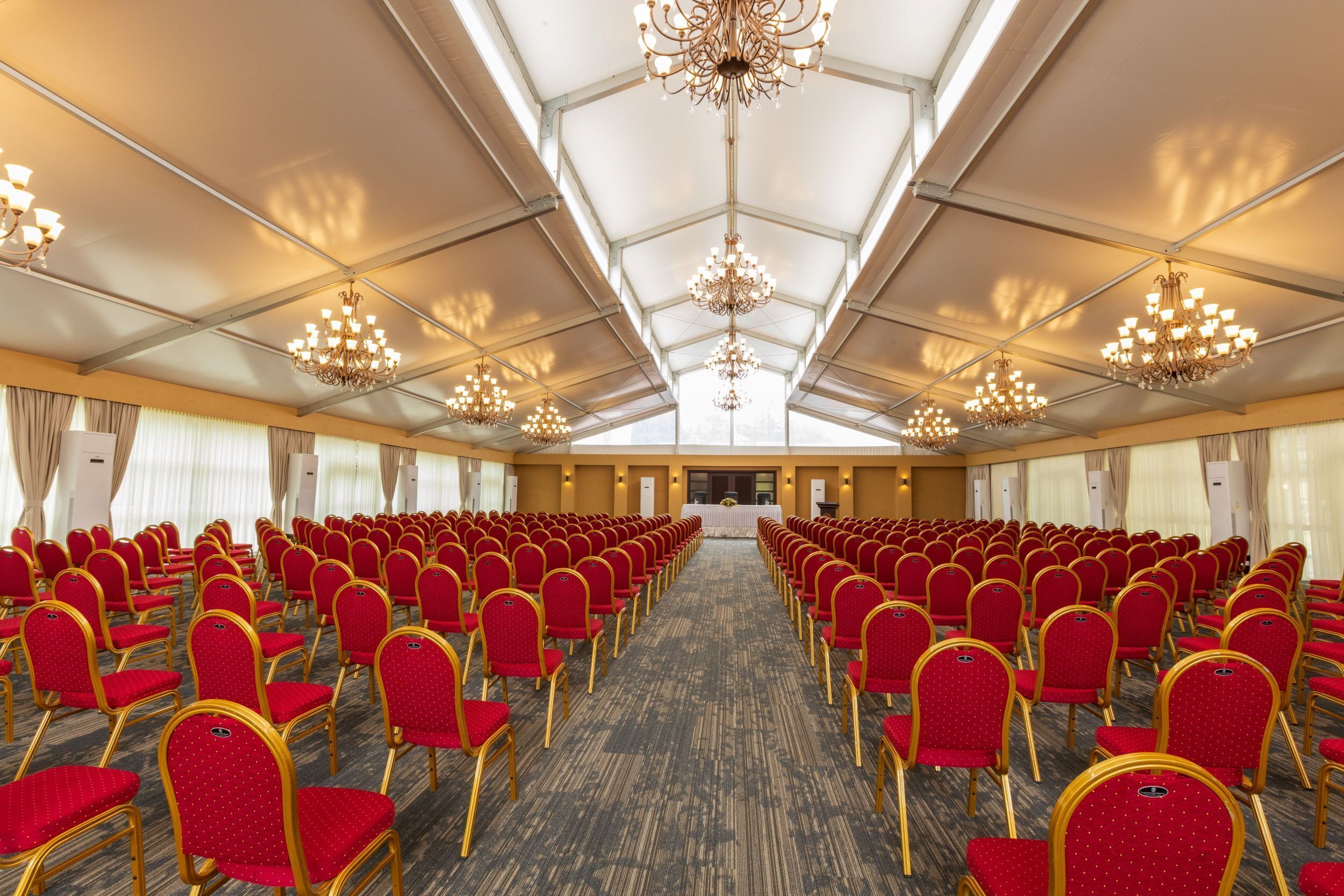 A conference room with rows of red chairs and gold frames, a carpeted floor, and chandeliers.