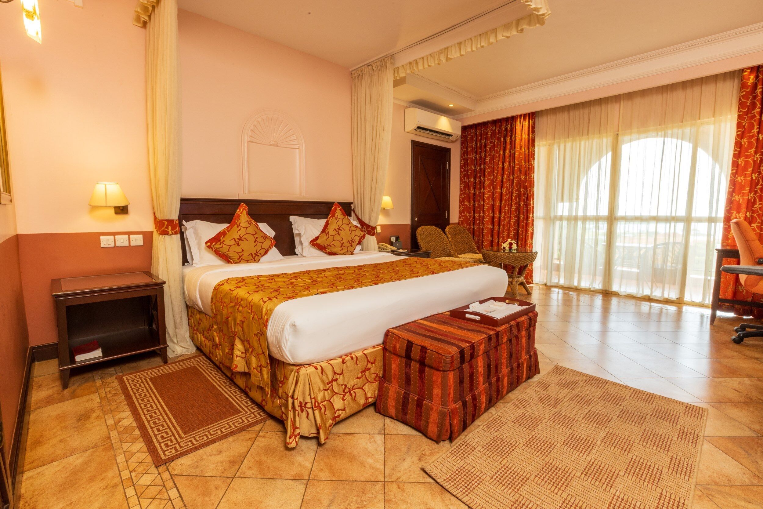 A spacious hotel room with a large bed, a wooden nightstand, a small table, and a seating area with a chair.