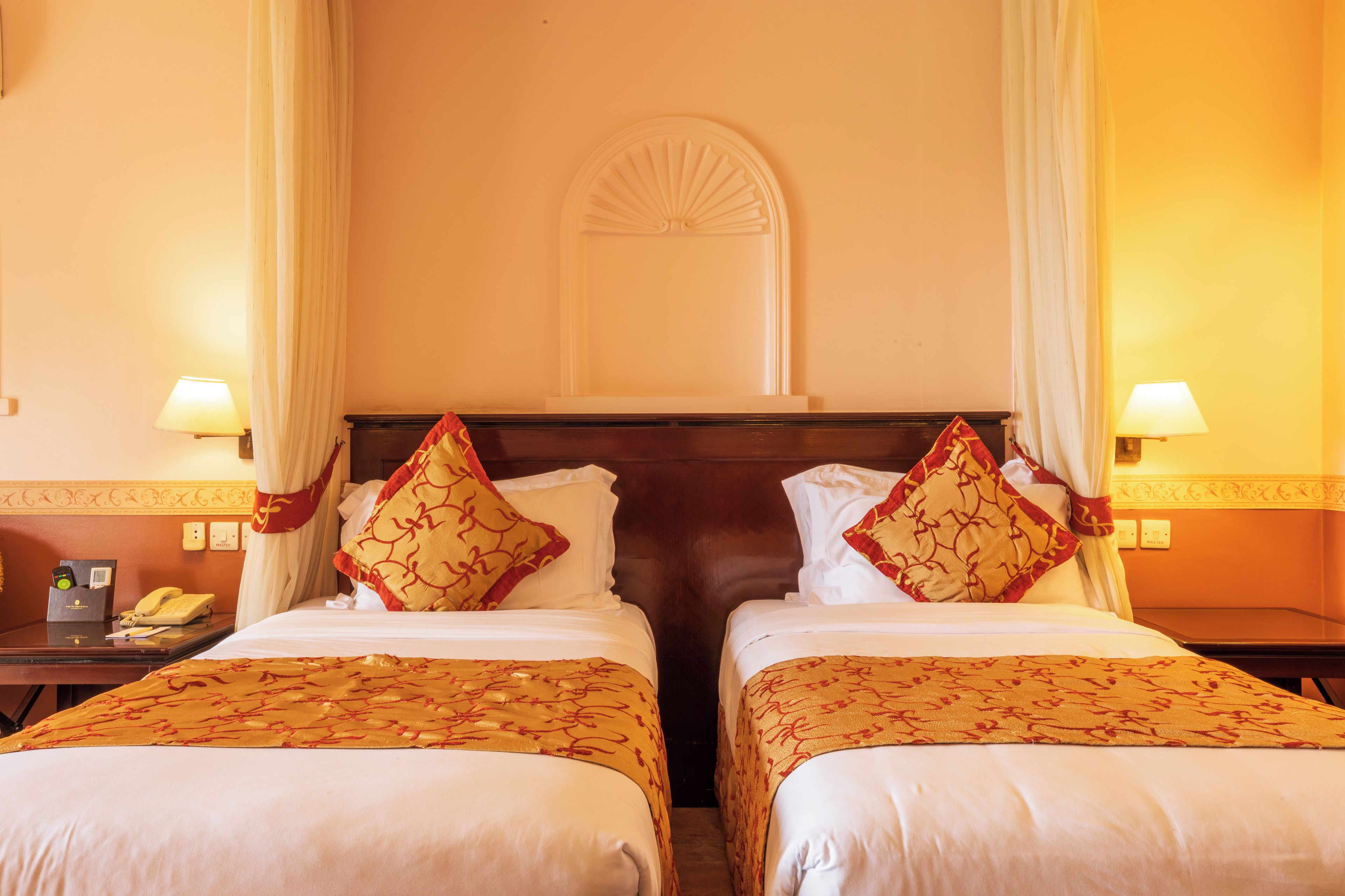 A hotel room with two beds, each with a white bedspread and red patterned pillows. There are two bedside tables with lamps, a telephone, and a minibar. The room has cream-colored walls and a decorative ceiling medallion.