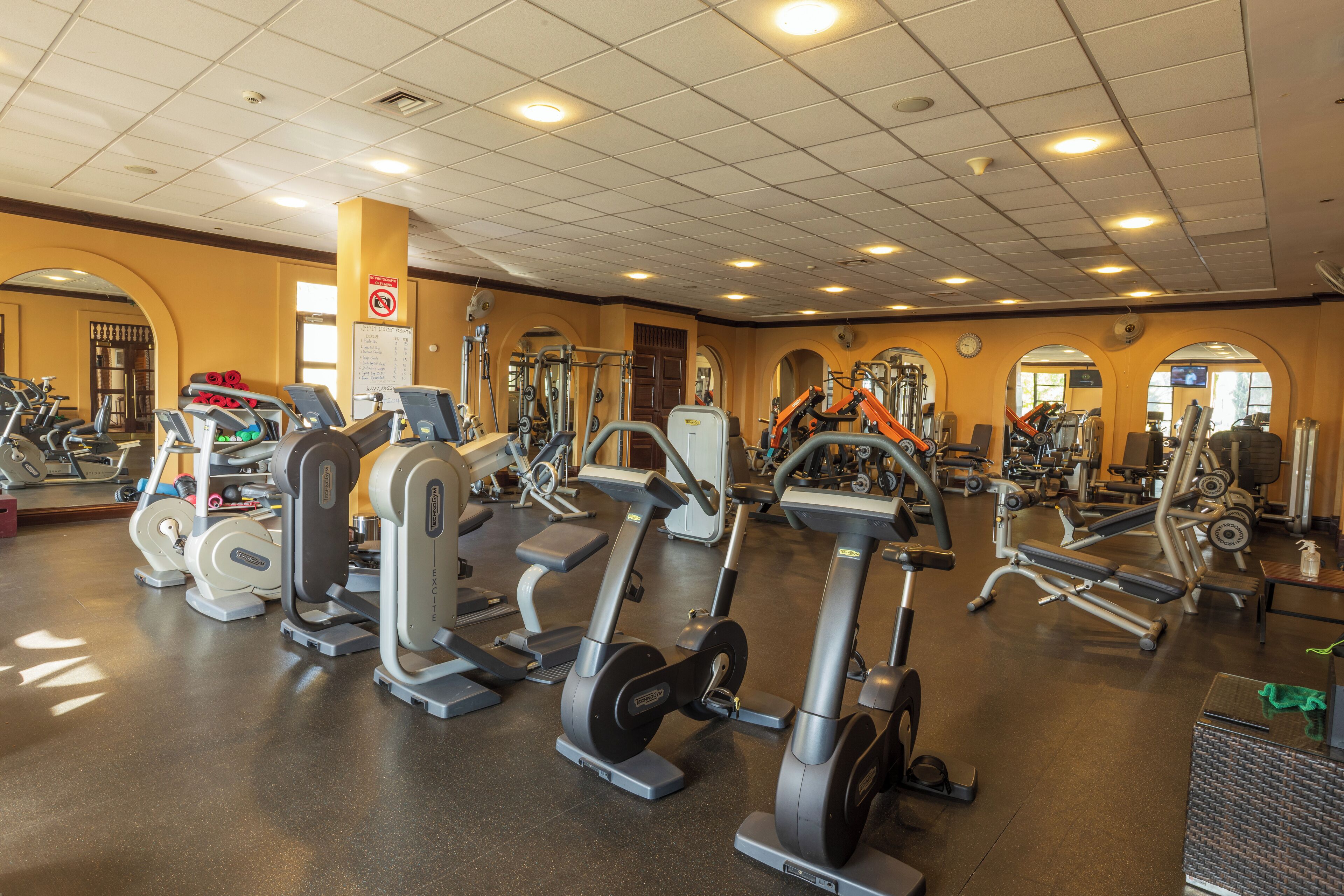 Ein Fitnessraum mit verschiedenen Trainingsgeräten, einer Tafel mit einer Liste und einer Uhr an der Wand.