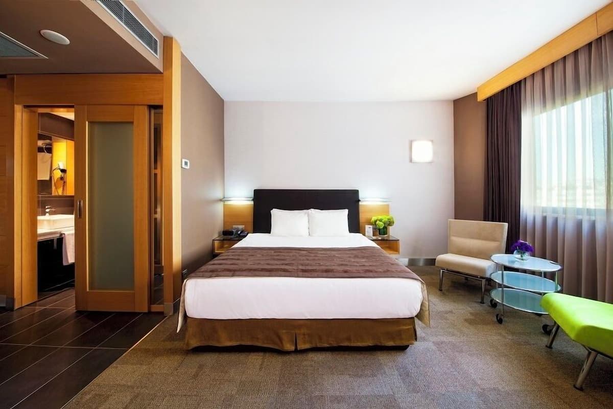 A hotel room with a large bed, bedside tables, a chair, a small table, and a bench.