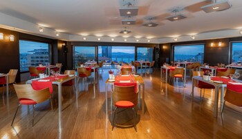 A spacious dining area with multiple tables and chairs, large windows offering a city view, and modern lighting fixtures.