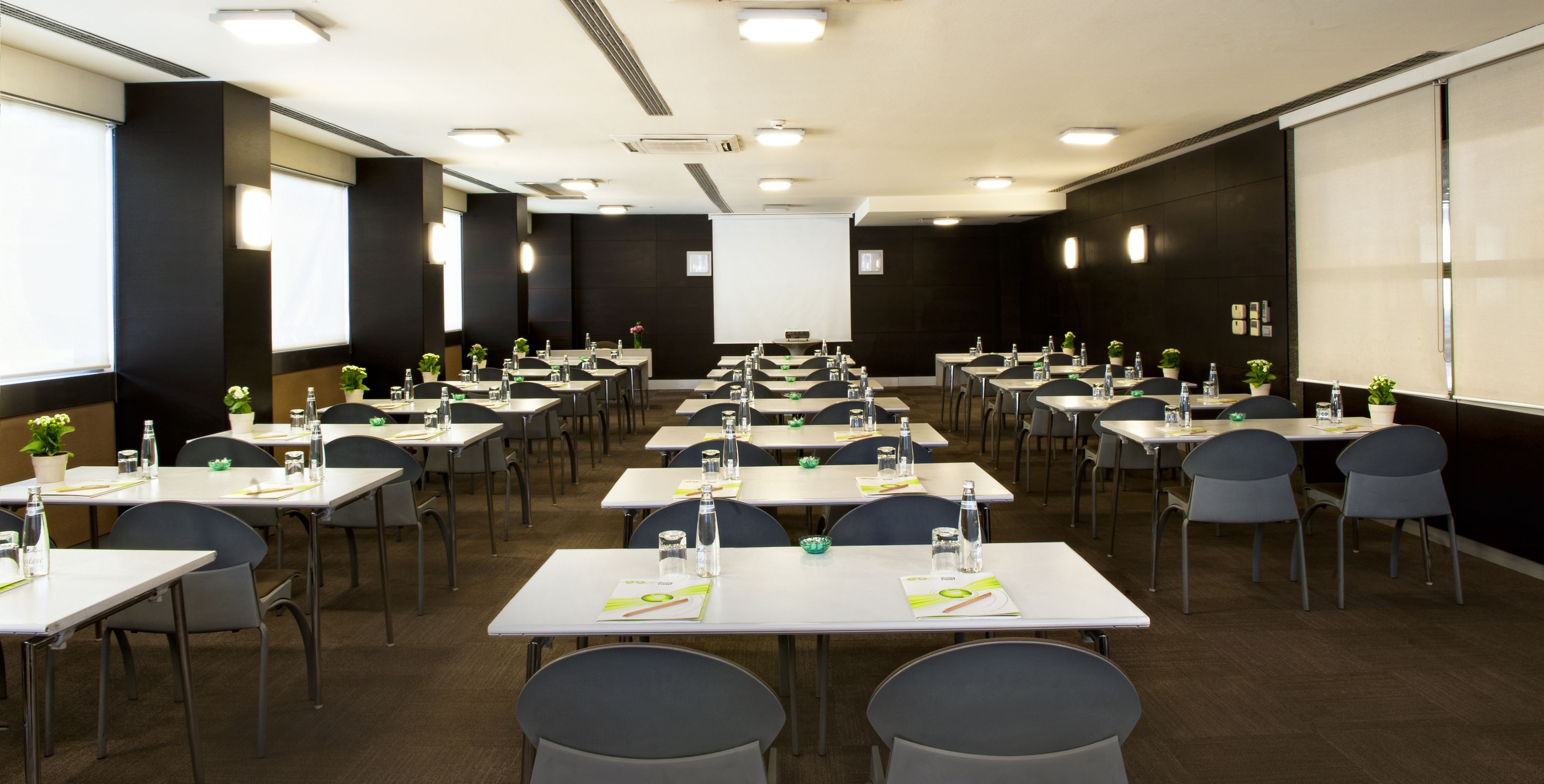 Un salón de conferencias con varias mesas, sillas y una pantalla de proyector.