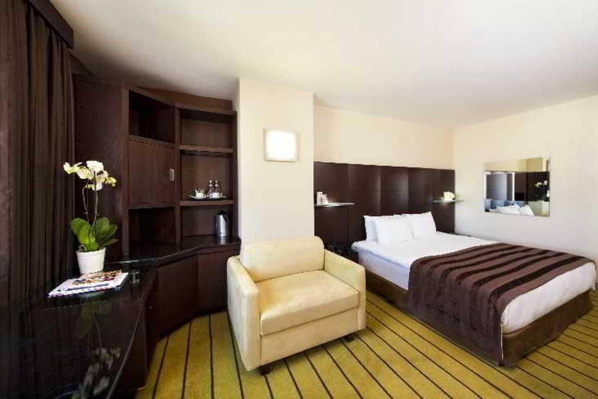 A hotel room with a bed, a chair, a wooden wardrobe, a small table with a plant, and a mirror on the wall.