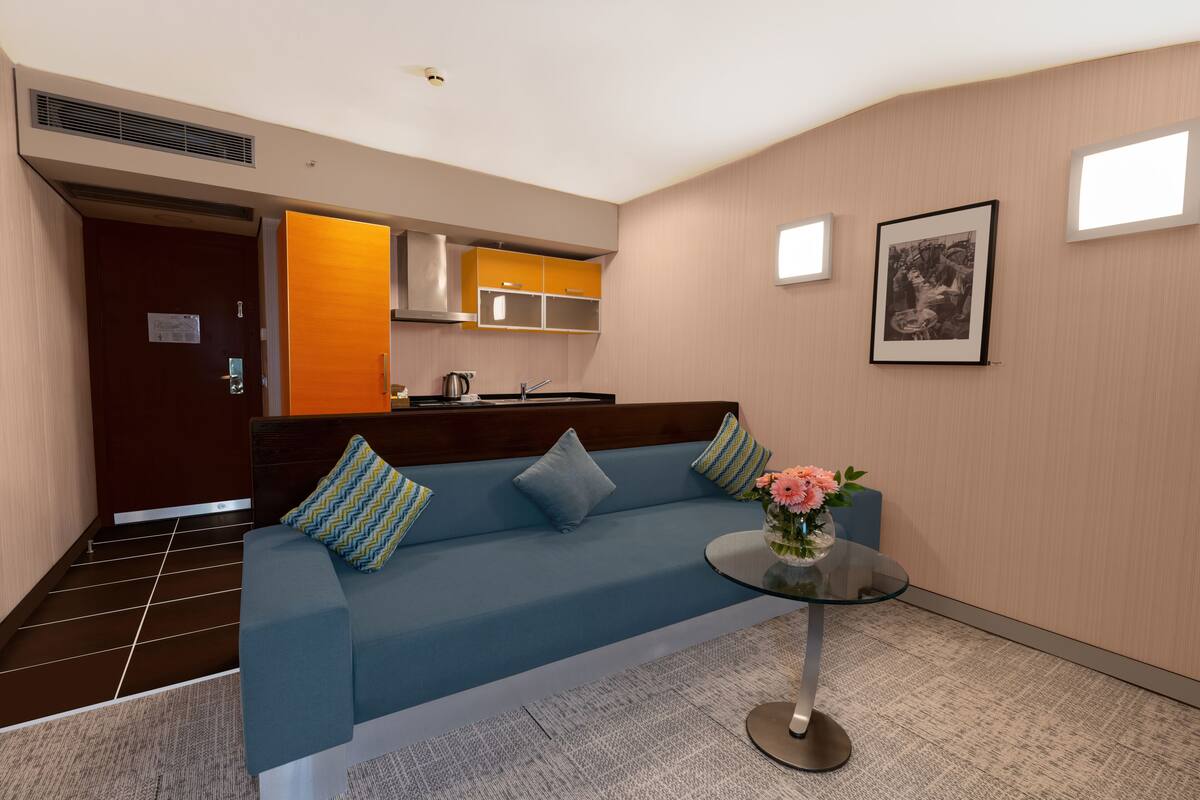 A modern hotel room with a blue sofa, a glass coffee table, and a kitchenette in the background.