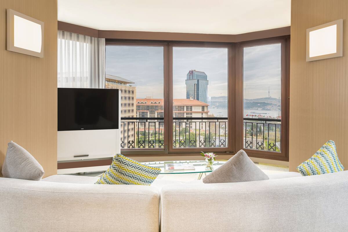 A modern living room with a large flat-screen TV, a sofa with patterned cushions, a coffee table with a vase, and a city view through large windows.