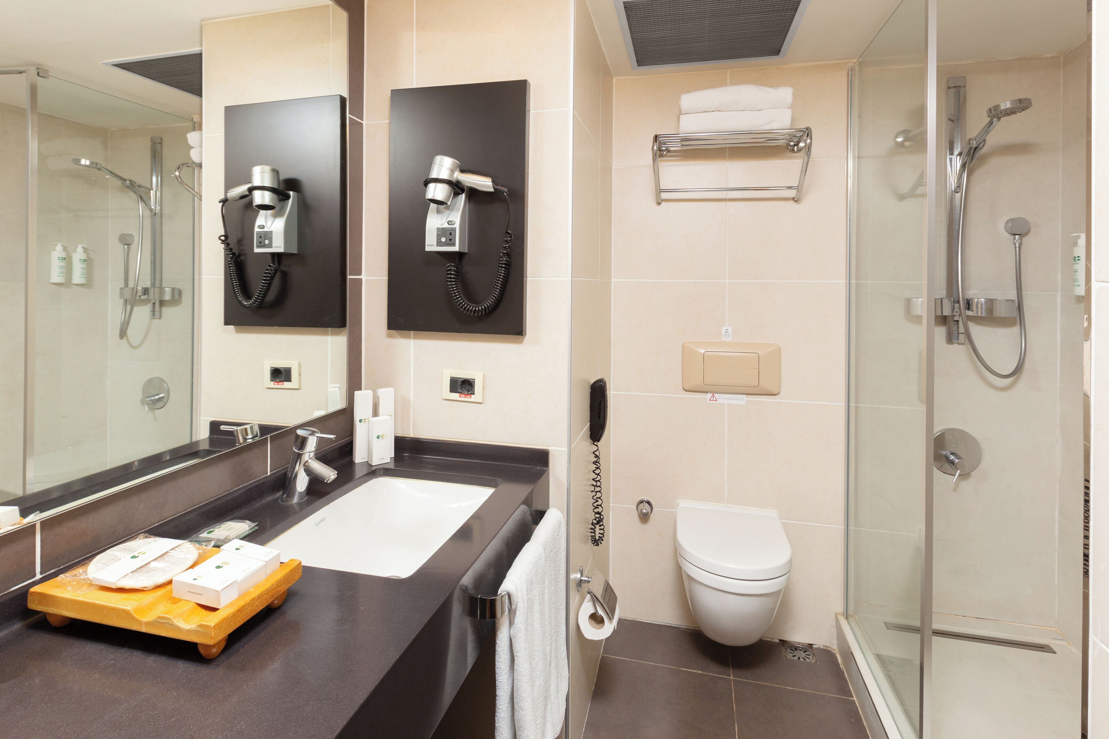 Un baño moderno con un espejo grande, un secador de pelo montado en la pared, un inodoro, una zona de ducha y un lavamanos con un perchero para toallas.
