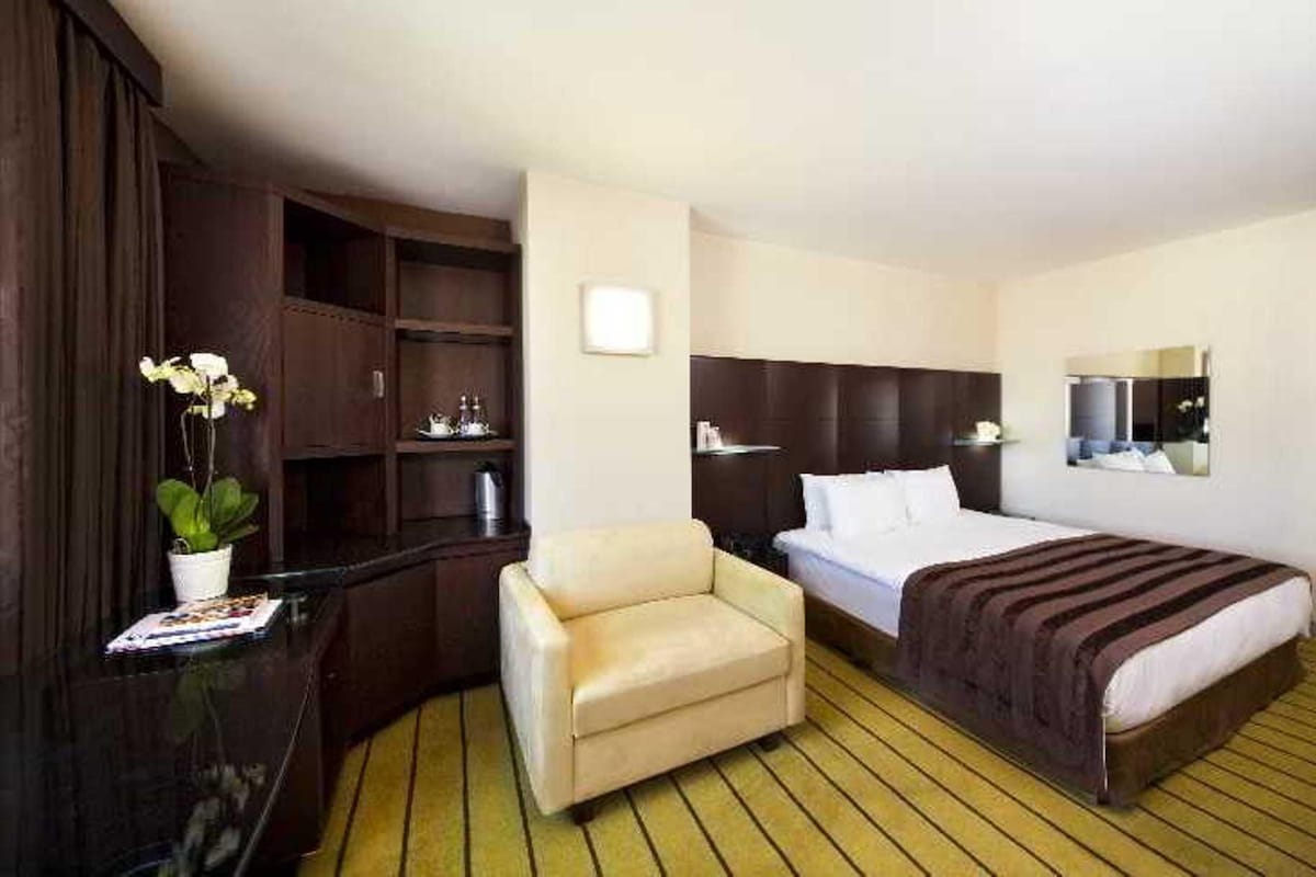 A hotel room with a bed, a chair, a wooden wardrobe, a small table with a plant, and a mirror on the wall.